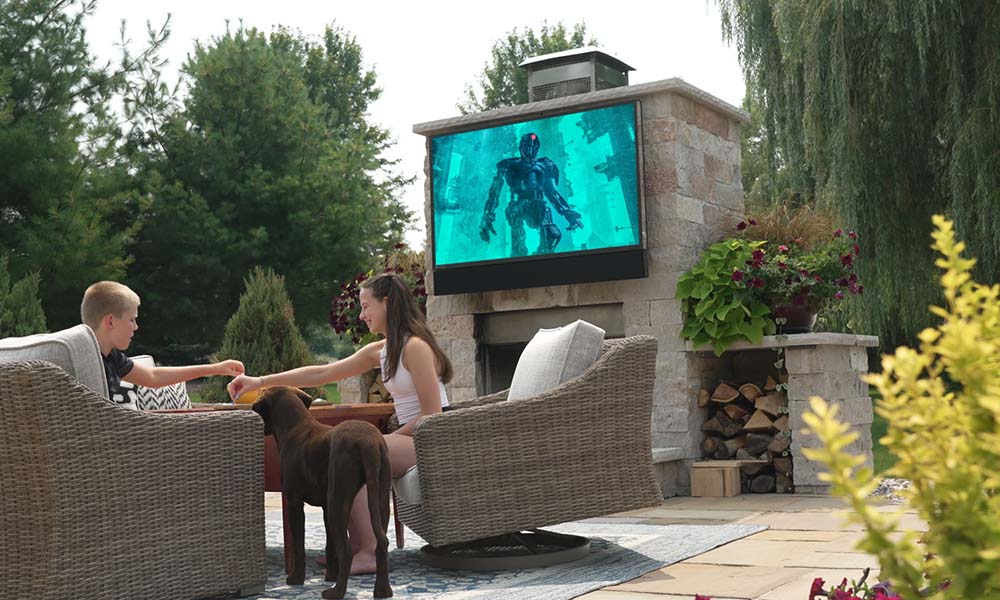 Two children sitting on outdoor wicker chairs with a dog nearby, watching a sci-fi movie on a large TV mounted above a stone fireplace in a landscaped backyard.
