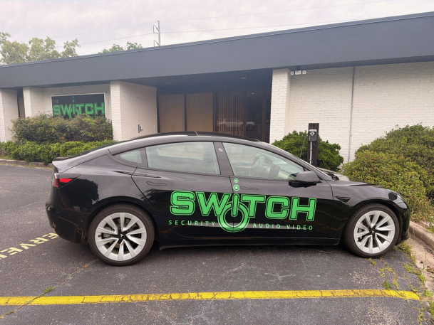 Black Tesla sedan with a green "SWITCH" logo parked along a yellow-striped parking space outside an office building.