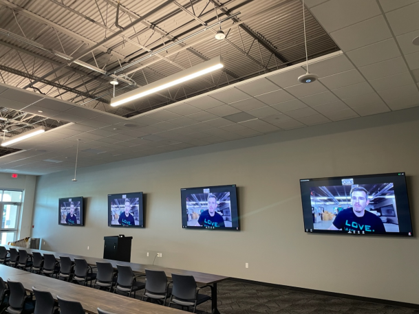 Large training room or auditorium featuring multiple wall-mounted screens displaying video feeds, long rows of dark tables, office chairs, and industrial exposed-ceiling lighting.