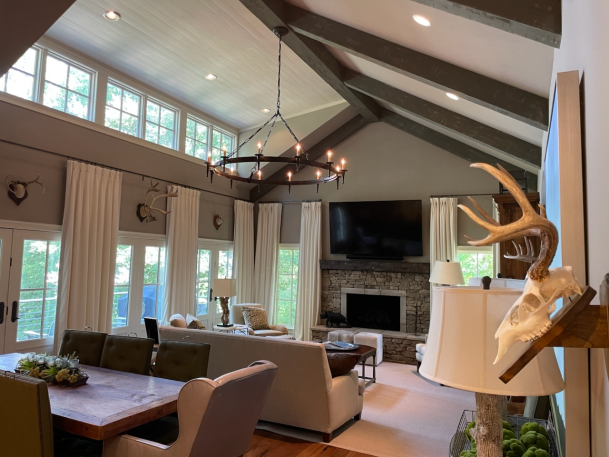 Open-concept living and dining area with vaulted dark-beamed ceilings, a large chandelier, and a stone fireplace.