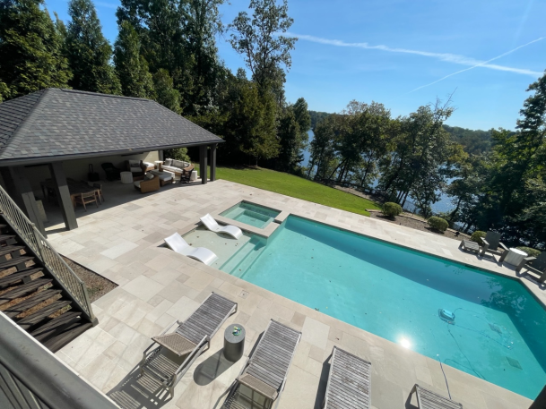 High-angle view of a luxury outdoor swimming pool with lounge chairs on a stone patio overlooking a lake surrounded by trees.