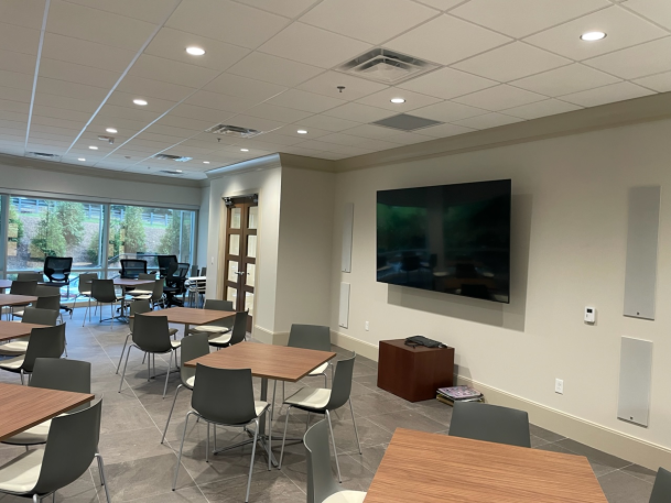 Large multipurpose room or cafeteria with several square dining tables, modern chairs, flush wall-mounted speakers, and a large central TV screen.