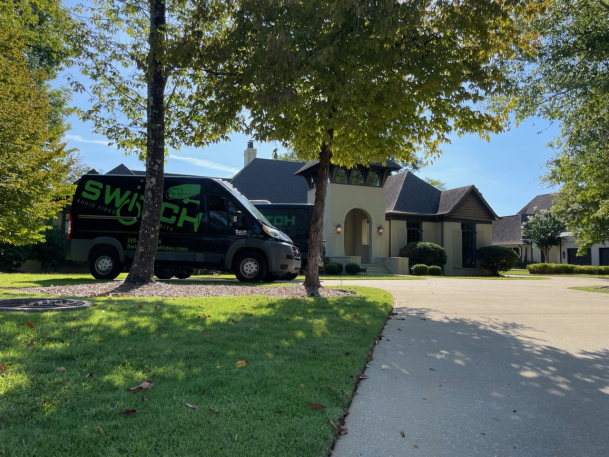 Black service van parked on a suburban street framed by shade trees in front of a residential house.