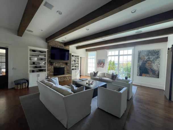 Living room with rustic dark wood ceiling beams, neutral sofas, large windows, and a built-in bookshelf.