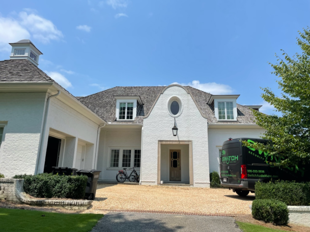 Exterior front view of a white brick storybook-style house with a gravel driveway and a black service van parked to the side.