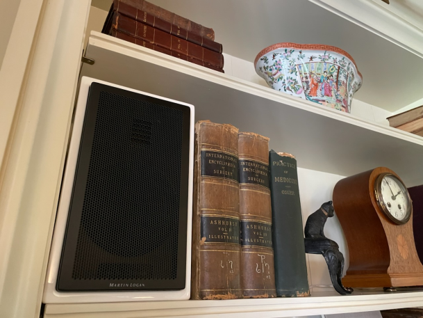 Close-up of a white bookshelf holding an integrated black audio speaker alongside vintage leather-bound books and a wooden clock.